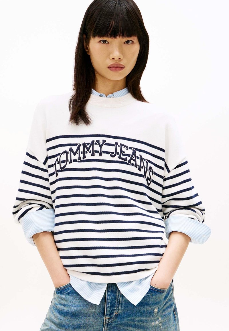Jeune femme aux cheveux raides et foncés portant un pull rayé blanc et bleu marine avec le texte "TOMMY JEANS", une chemise bleu clair et un jean bleu, les mains dans les poches.