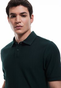 Ciemnozielona, prążkowana koszulka polo z usztywnionym kołnierzykiem, z dwoma guzikami przy dekolcie. Materiał wydaje się mieć fakturę i jest lekki.