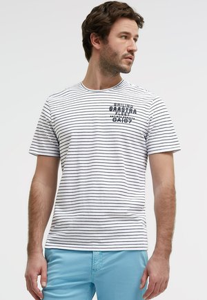 Mann trägt weißes T-Shirt mit dünnen schwarzen horizontalen Streifen und marineblauem Text, kombiniert mit hellblauen Hosen, schaut zur Seite.