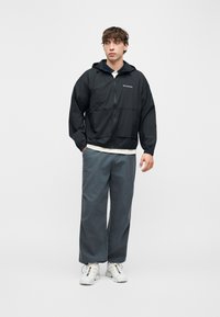 Νέος άνδρας που στέκεται, φοράει μαύρο μπουφάν Columbia με φερμουάρ, γκρι παντελόνι φαρδύ, λευκά αθλητικά παπούτσια, με τα χέρια χαλαρά, σε απλό φωτεινό φόντο.