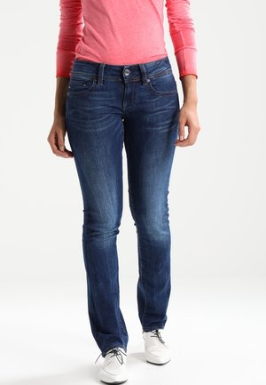 Frau trägt dunkelblaue, enganliegende Jeans, korallenrotes Langarmshirt und weiße Schnürschuhe, steht vor einem schlichten Hintergrund.