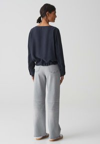 Blusa blu navy a maniche lunghe con una vestibilità ampia; scollatura rotonda e orlo arricciato, abbinata a jeans grigi chiari a gamba larga e scarpe casual.