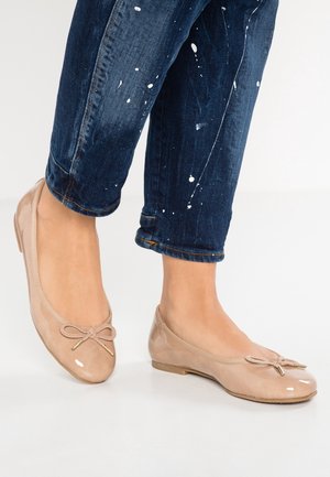 Beige ballet flats i laklæder med sløjfedetalje, båret med mørkeblå bukser med malingstænk på en lys gulv.