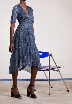 Femme portant une robe midi à couches bleue à motifs avec des manches aux coudes, des sandales à talons carrés noires, debout à côté d'une chaise moderne bleue.