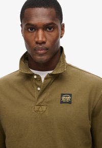 Khakigrünes Langarm-Polohemd mit umgeschlagenem Kragen, einer Drei-Knopf-Leiste und einem Logopatch auf der linken Brust. Strukturierter Stoff.