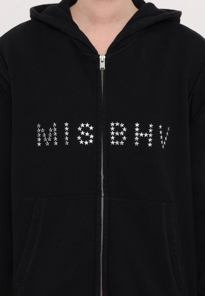 Hanorac negru cu fermoar pe față, două buzunare și lettering cu model de stea argintie care scrie "MISBHV." Material moale și croială lejeră.