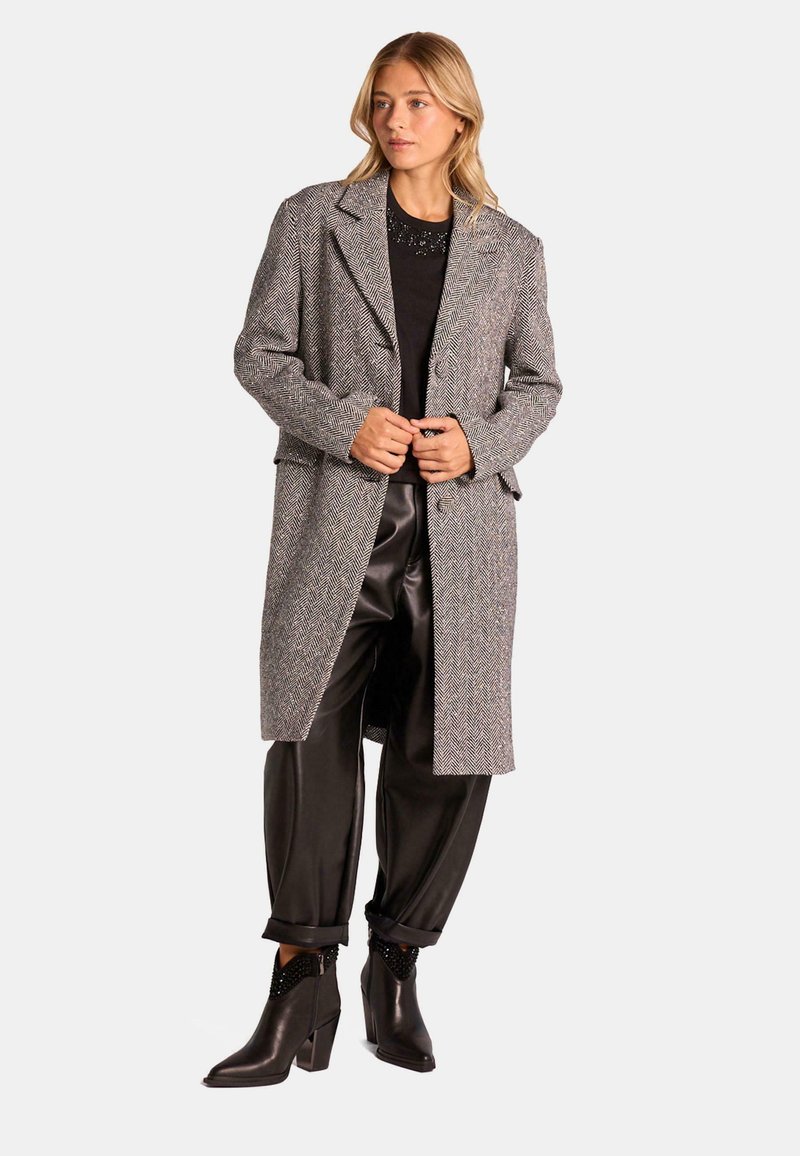 Abrigo gris de espiga con cuello, mangas largas y abertura frontal, combinado con pantalones de satén negros y botines que presentan acentos texturizados.