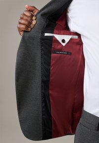 Szary blazer z elastycznej tkaniny, z bordowym satynowym wykończeniem, z czarną kieszenią i metką z napisem "najlepsza elastyczna tkanina".