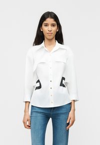 Γυναίκα με μακριά σκούρα μαλλιά που φοράει λευκό πουκάμισο με κουμπιά και μαύρες λεπτομέρειες, καθώς και μπλε τζιν, στέκεται μπροστά σε απλό φωτεινό φόντο.