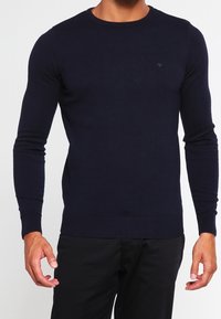 Pull tricoté bleu marine avec col rond, manches longues et poignets et ourlet côtelés. Texture lisse avec un petit logo en détail.