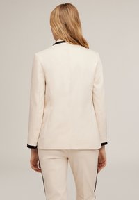 Blazer crème avec une coupe ajustée, bord noir sur les poignets et texture lisse. L'arrière présente des lignes épurées et un design structuré.
