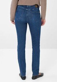 Jean en denim bleu avec une coupe droite, présentant des poches arrière et une étiquette en cuir marron. Le tissu a une texture lisse avec une décoloration minimale.