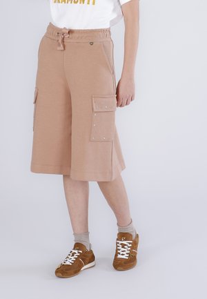 Persona che indossa pantaloncini cargo beige fino al ginocchio con tasche borchiate, una t-shirt bianca, calzini beige e sneakers marroni con lacci bianchi.
