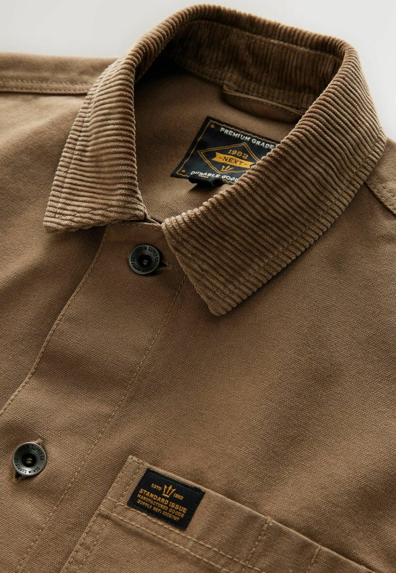 Brown shirt with a corduroy collar, featuring a button closure, fabric tag, and a textured finish. Key detail includes a small patch near the hem.