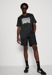 Black Adidas Originals t-shirt with a diagonal pattern and large white logo, paired with black shorts and black sneakers with white stripes.