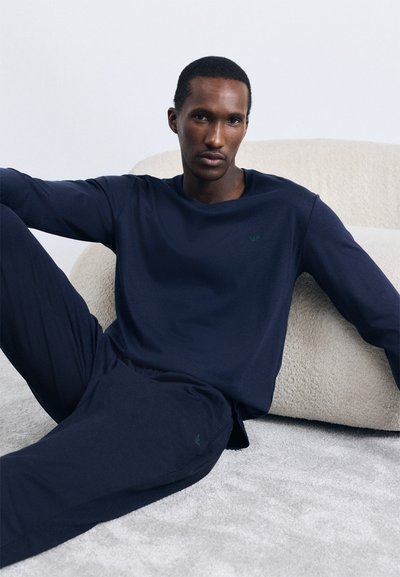 Navy blue loungemekko, joka on valmistettu pehmeästä kankaasta; siinä on pitkät hihat, löysät housut ja hillitty logoyksityiskohta rinnassa.