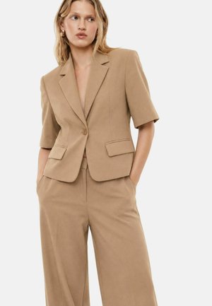 Donna che indossa un blazer color cammello a maniche corte e pantaloni ampi coordinati, in piedi con le mani nelle tasche su uno sfondo bianco semplice.