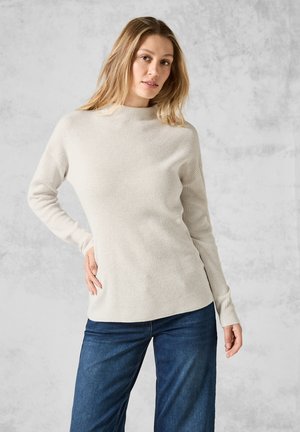 Ribba, krämfärgad, långärmad tröja med hög halsringning. Buren med blå jeans som har en figursydd design och subtil textur.