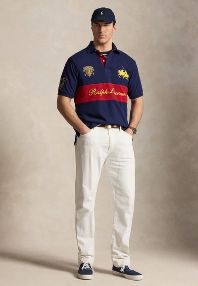 Άνδρας που φορεί μπλε σκούρο και κόκκινο πόλο Ralph Lauren, άσπρο παντελόνι, μπλε σκουφάκι και μπλε αθλητικά, στέκεται με τα χέρια στις τσέπες.