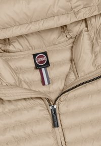 Beige pufferjack met ritssluiting, horizontale stiksels en een merklabel. Heeft een glad, glanzend oppervlak en contrasterende accentstrepen.