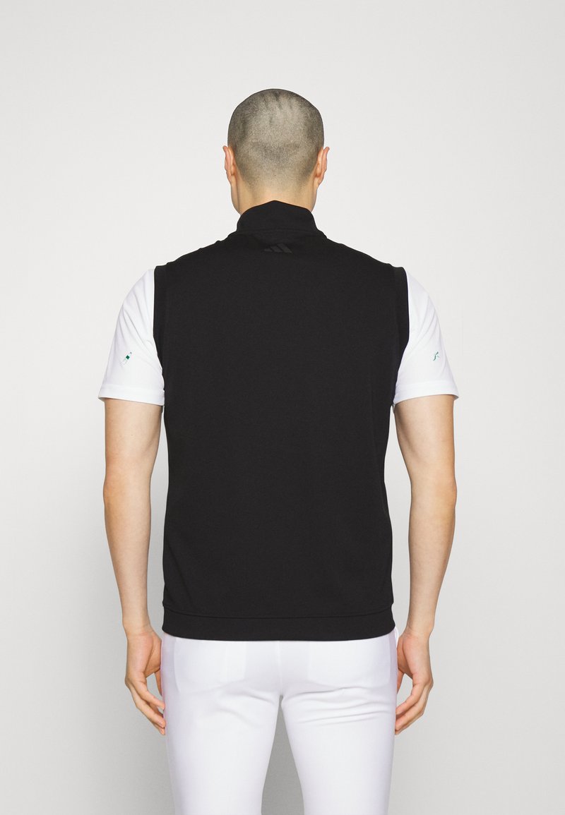 Zwarte mouwloze vest met een hoge kraag, een gladde textuur en een getailleerd ontwerp, gedragen over een wit T-shirt met korte mouwen. Subtiele branding zichtbaar.