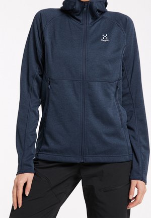 Personne portant une veste bleu marine zippée avec capuche et poches zippées, associée à un pantalon noir.