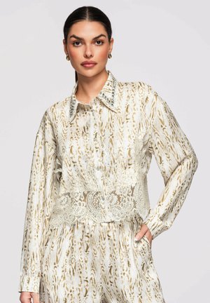 Femme aux cheveux foncés porte un chemisier beige et blanc à manches longues avec un détail en dentelle et un pantalon assorti, posant avec une main dans la poche.