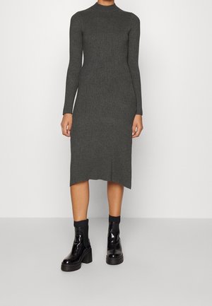 Kvinde iført en mørkegrå, ribstrikket midi-kjole med lange ærmer, kombineret med sorte chunky platform-ankelstøvler mod en ensfarvet baggrund.