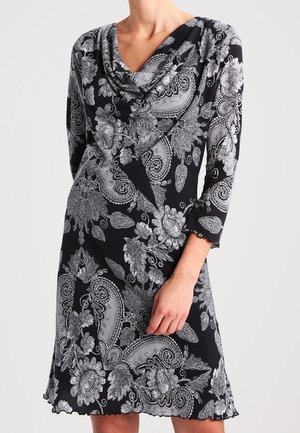 Schwarzes Kleid mit weißen Blumen- und Paisley-Mustern, mit einem drapierten Ausschnitt und Dreiviertelärmeln, getragen von einer Person.