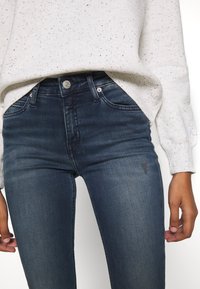 Persona con jeans skinny de color azul oscuro con un ligero desgaste y un suéter claro con motas, con las manos relajadas a los lados.