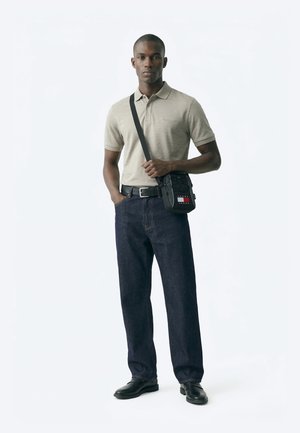 Man draagt beige poloshirt, donkere spijkerbroek, zwarte riem, zwarte schoenen en een zwarte crossbodytas met Tommy Jeans-logo, staand tegen een witte achtergrond.