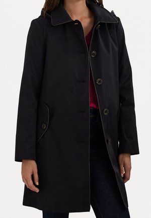 Femme portant un manteau noir boutonné longueur genoux avec col, par-dessus un haut rouge foncé et un pantalon sombre, debout, bras détendus le long du corps.