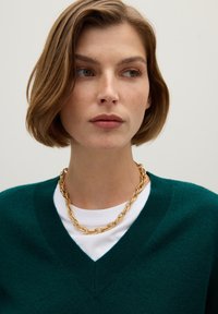 Suéter verde de pico sobre una camisa blanca, acompañado de un collar de cadena dorada. Textura suave, ajuste ceñido y colores contrastantes.