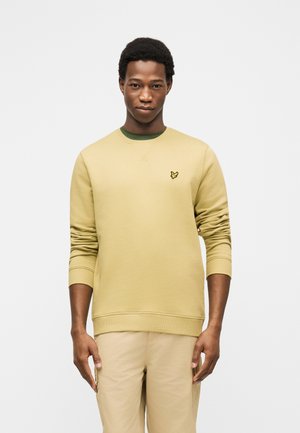 Jonge man met een beige sweatshirt met een klein vogel-logo op de borst, gecombineerd met lichtbeige broek, staand tegen een effen achtergrond.