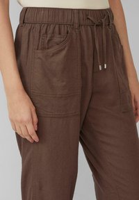 Pantalons légers marron avec une taille élastique, un cordon de serrage et plusieurs poches. Le tissu présente une finition douce et texturée.
