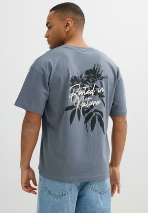 Hombre con camiseta gris con diseño floral negro y texto "Rooted in Nature" en la espalda, combinado con vaqueros denim azul claro.