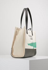 Borsa tote in tela con manici in pelle sintetica nera, grafica di un alligatore verde e testo, caratterizzata da una parte superiore crema e una inferiore grigia.
