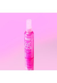 Krulverfrissingsspray in een transparante roze fles met een witte spray-nozzle. Het etiket toont tekst in wit en roze. Reflecterend oppervlak eronder.