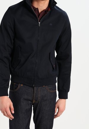 Homme portant une veste noire zippée sur une chemise rouge foncé associée à un jean bleu foncé, sur un fond blanc uni.