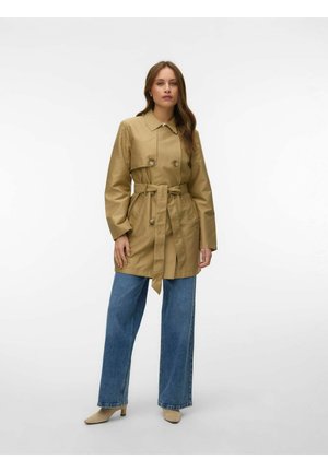 Donna con trench beige con cintura, jeans blu a gamba larga e scarpe beige con tacco, in piedi su uno sfondo bianco semplice.