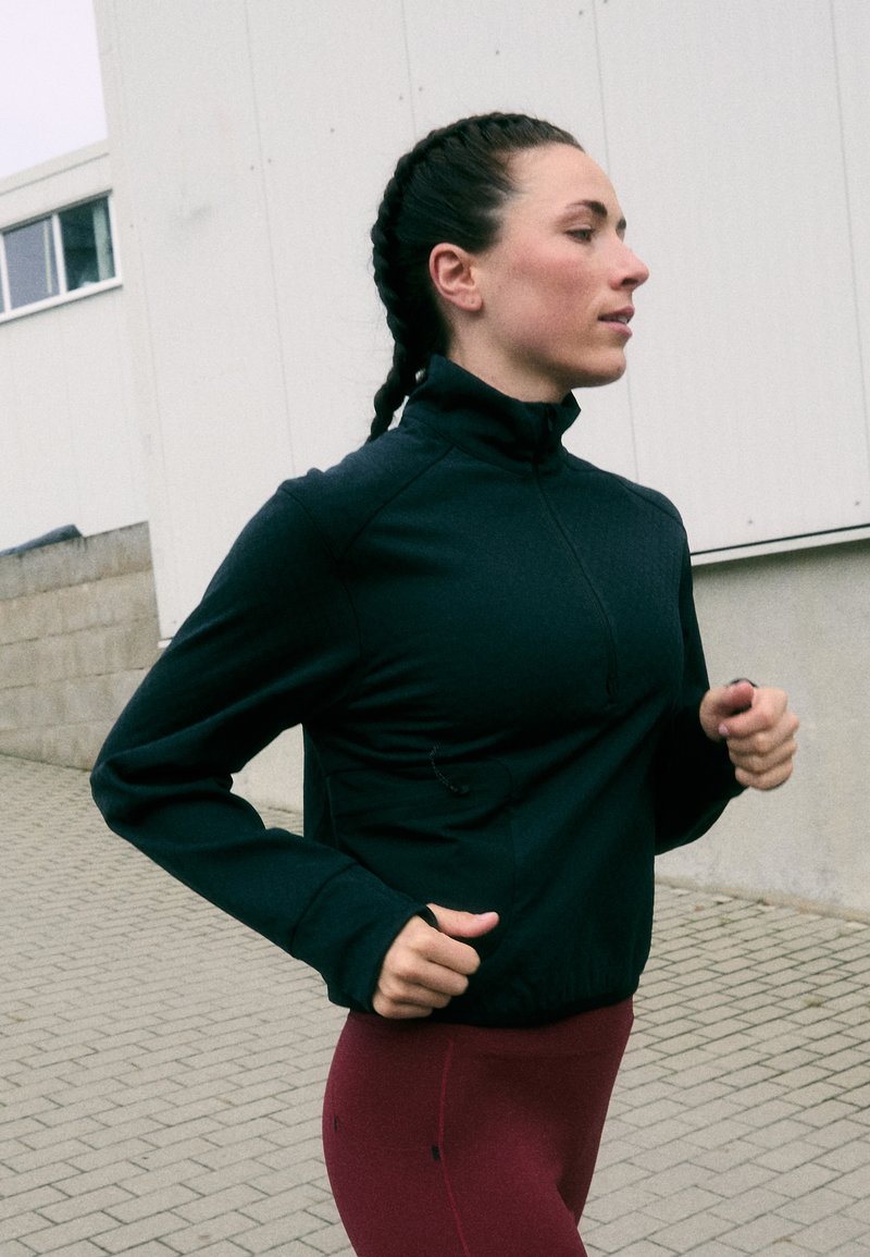 Črna atletksa jopa z zadrgo, visokim ovratnikom, oprijeto zasnovo in teksturirano tkanino, kombinirana s temnordečimi pajkicami, na tlakovanem zunanjem površju.