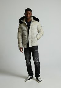 Puffete lys beige jakke med hette av svart kunstpels, glidelås foran og teksturert søm, kombinert med slitte mørkejeans og sneakers.