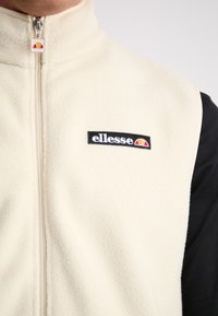 Krem krāsas vilnas veste ar melnu logotipa uzlīmi. Aprīkota ar augstu apkakli un rāvējslēdzēja aizdari priekšpusē. Tekstūrēta auduma un kontrastējošas piedurknes detaļa.