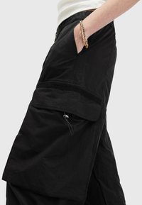 Personne portant un pantalon cargo noir avec poches latérales, main dans la poche, et un bracelet chaîne doré au poignet sur un fond uni.