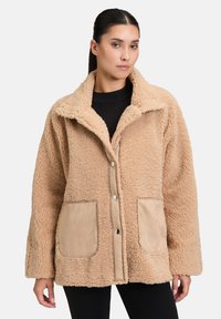 Veste en fausse fourrure beige avec un col structuré, deux poches avant en suède et fermeture à boutons-pression. Tissu doux et texturé.