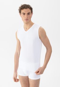 Jonge man in een mouwloze witte onderhemd en witte boxershort, die de zoom van het shirt vasthoudt, tegen een effen witte achtergrond.