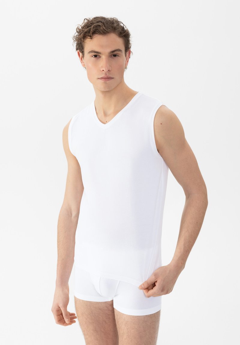 Jonge man in een mouwloze witte onderhemd en witte boxershort, die de zoom van het shirt vasthoudt, tegen een effen witte achtergrond.