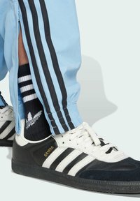 Black and white Adidas Samba sneakers with a textured sole next to light blue track pants featuring black stripes and zippered hems.