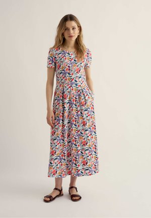 Žena stojaca v krátkom rukáve, šaty po členky s farebnými kvetinovými vzormi a hnedé sandále s odkrytými prstami na svetlom pozadí.