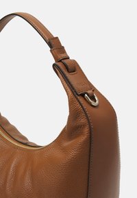 Brun lædertaske med teksturerede og glatte paneler, som har en lille metalring og broderede håndtagsdetaljer.